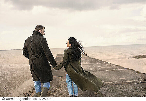 Couple in winter coats holding hands on ocean beach jetty