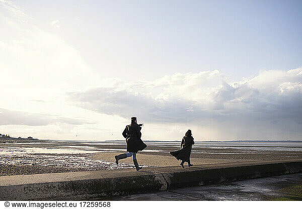 Couple in winter coats running on sunny ocean beach jetty