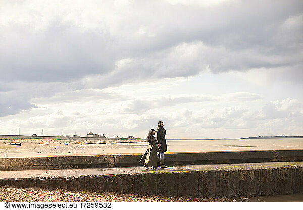 Couple in winter coats walking on sunny winter ocean beach jetty