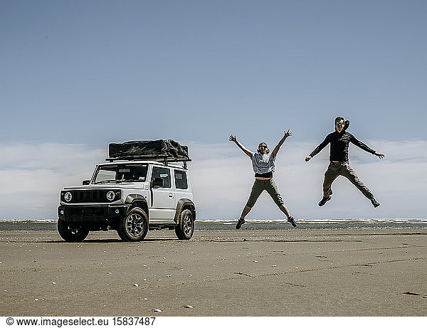couple jumps for joy next to their SUV parked on beach in New Zealand