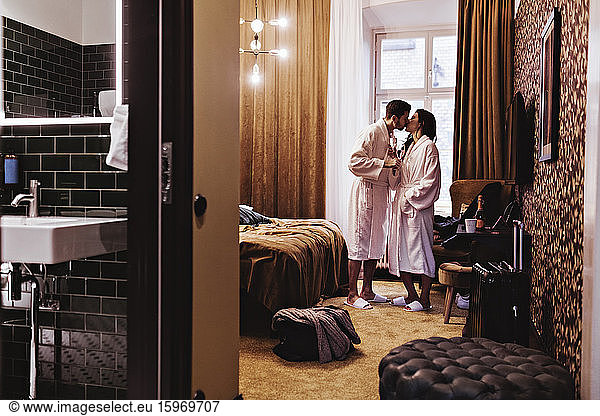Couple kissing while holding champagne flutes by bed in hotel room