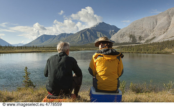 Couple  man and woman  sitting on the shore  enjoying panorama  view  Wind River Valley  Northern Mackenzie Mountains  Yukon Territory  Canada