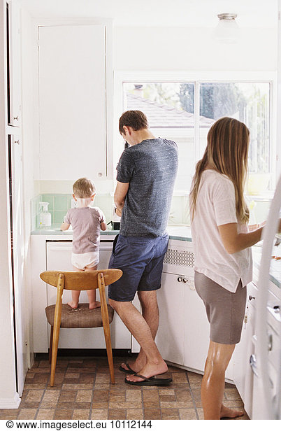 Couple standing in a kitchen  their son standing on a chair beside them.