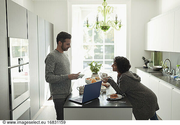 Couple talking and enjoying breakfast at laptop in morning kitchen