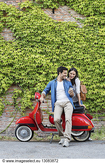 Couple using smart phone while standing with motor scooter against ivy wall