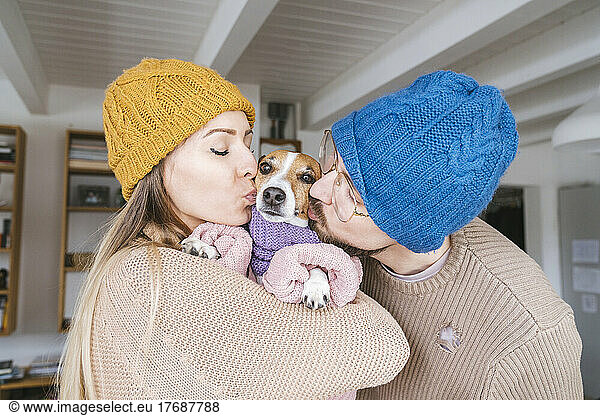 Couple wearing wooly hat at home kissng dog with scarf