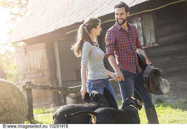 Couple with saddle and dogs holding hands outside rural barn