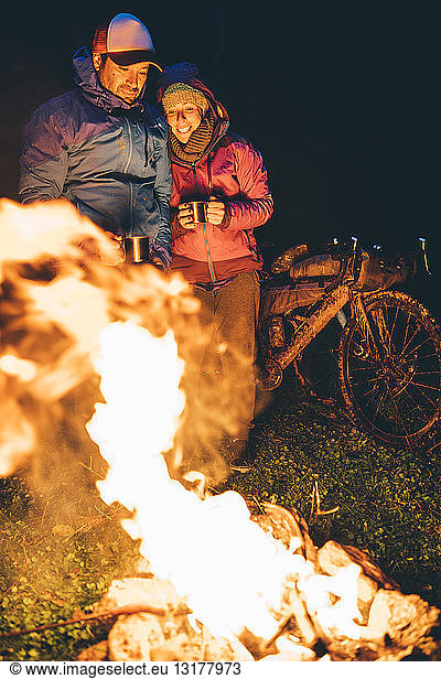 Couple with with bikes standing at camp fire looking at flame by night