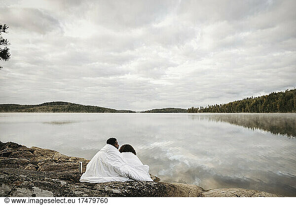 Couple wrapped in blanket sitting at lakeshore against cloudy sky
