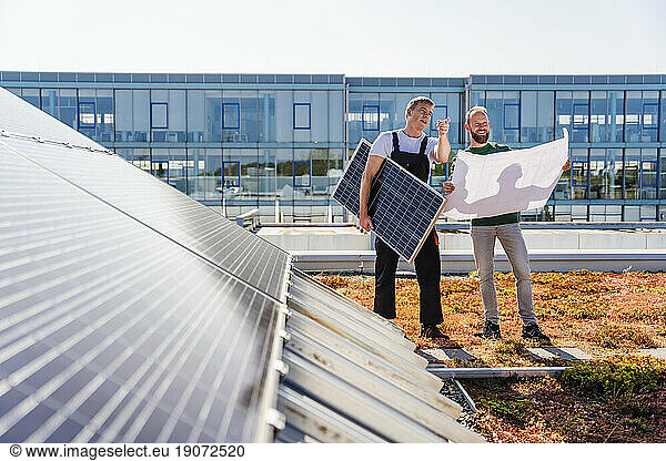Craftsman and colleague with plan and solar panel on the roof of a company building