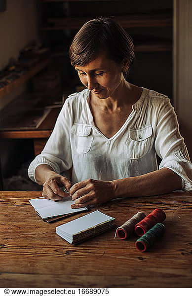 Craftswoman stitching sheets of paper for book  sitting at table