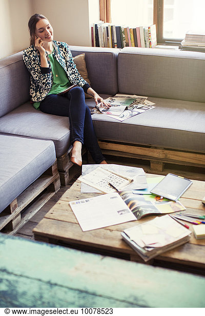 Creative businesswoman reviewing proofs and talking on cell phone on office sofa