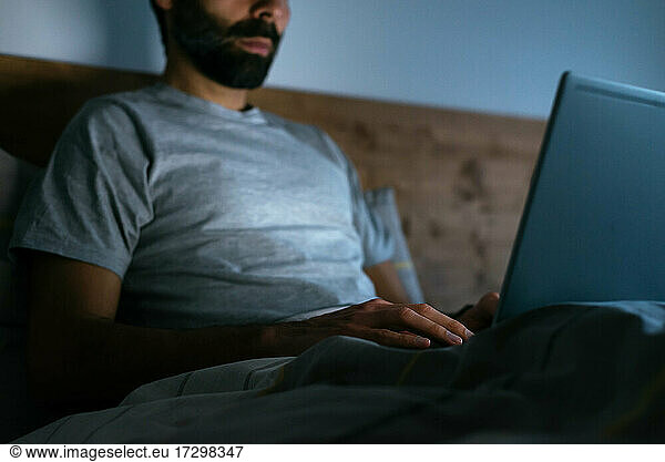 Crop man using a laptop on bed at night