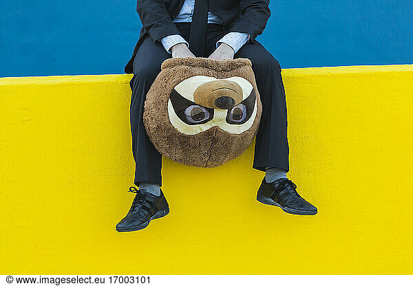 Crop view of businessman in black suit sitting on yellow wall holding with meerkat mask