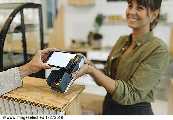 Cropped hand of customer doing contactless payment with smart phone to female owner at checkout in coffee shop