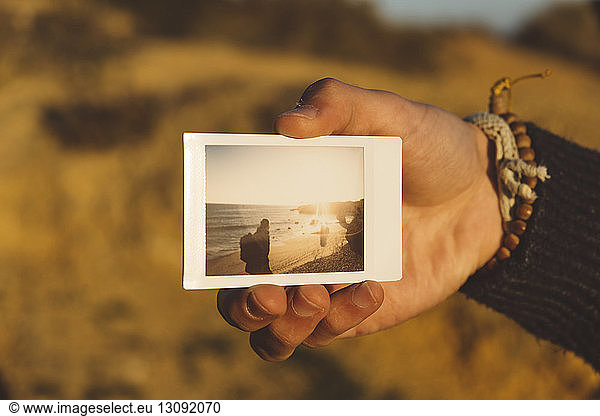 Cropped hand of man holding instant photograph at beach during sunset