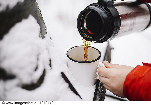 Cropped hands of woman pouring black coffee from insulated drink container into mug on snow