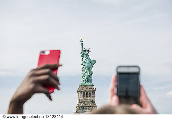 Cropped hands using smart phones against Statue of Liberty and cloudy sky in city