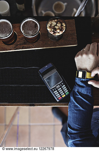 Cropped image of customer doing contactless payment through smart watch by beer glasses at bar counter