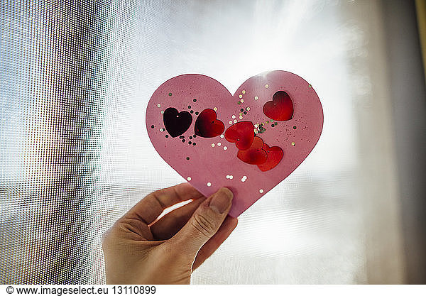 Cropped image of hand holding heart shape artwork against window
