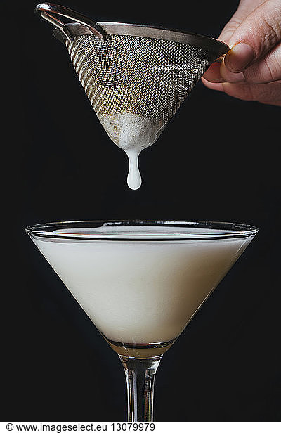 Cropped image of hand straining drink in martini glass against black background
