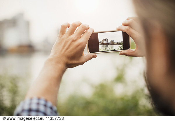 Cropped image of man photographing silo through smart phone against river
