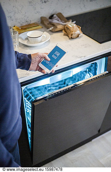 Cropped image of man using mobile app while standing by dishwasher at smart home