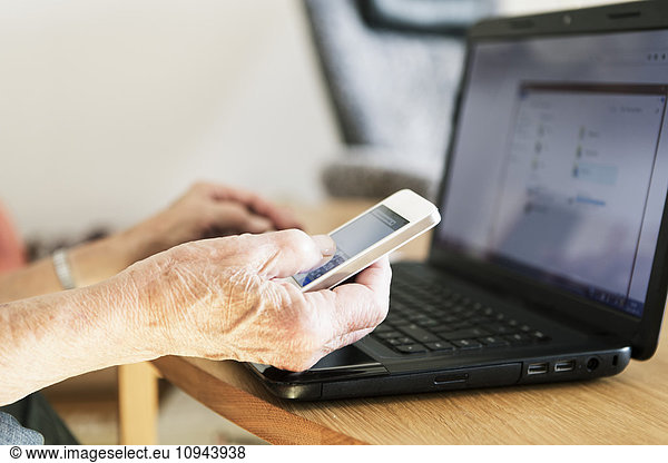 Cropped image of senior woman's hand using laptop and phone at home