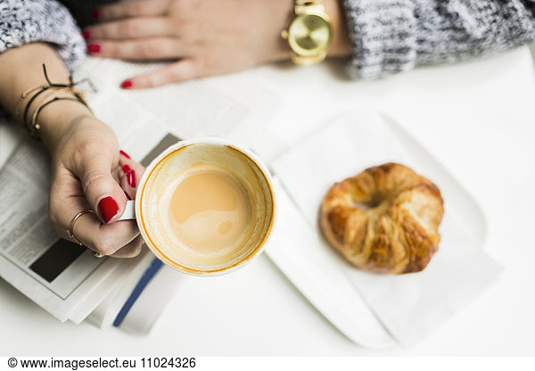 Cropped image of woman holding coffee cup over croissant and newspaper