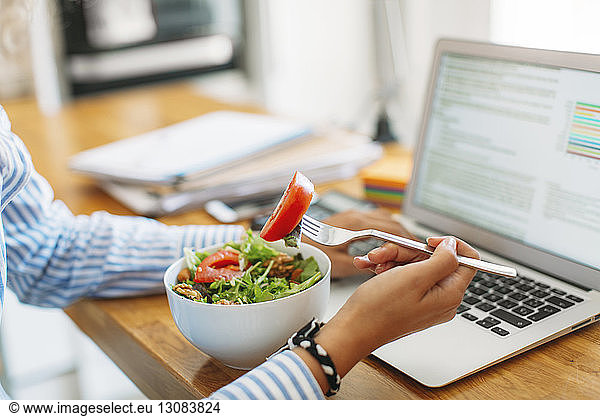 Cropped image of woman holding fork with tomato slice by laptop computer at wooden table
