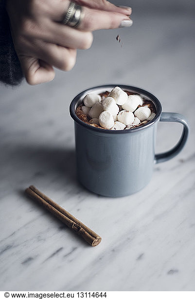 Cropped image of woman sprinkling cocoa powder on hot chocolate marshmallow mug