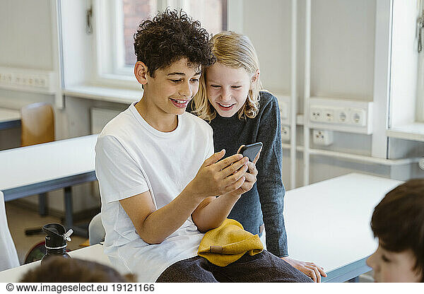 Curious boy and girl sharing smart phone while sitting at desk in classroom
