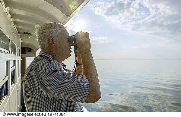 Curious senior man looking at sea through binoculars from ship