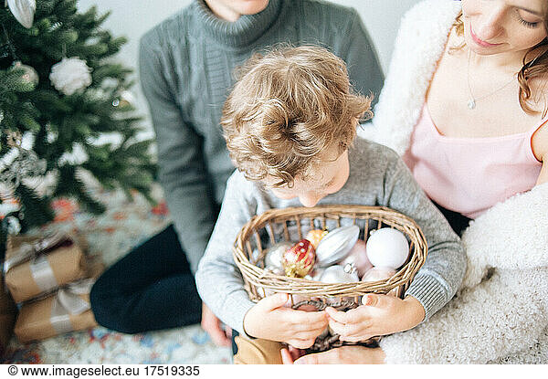 Curly boy hugs basket with Xmas decor while sitting on mother legs