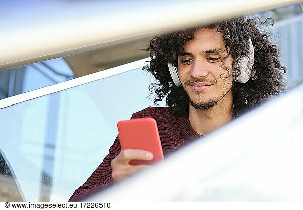 Curly haired young man using smart phone while listening music through headphones