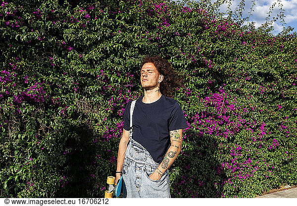 curly red hair tattoo men with skateboard against plants wall