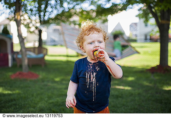 Cute baby boy eating ice cream while standing on grassy field at yard