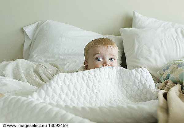 Cute baby boy hiding amidst duvet on bed at home