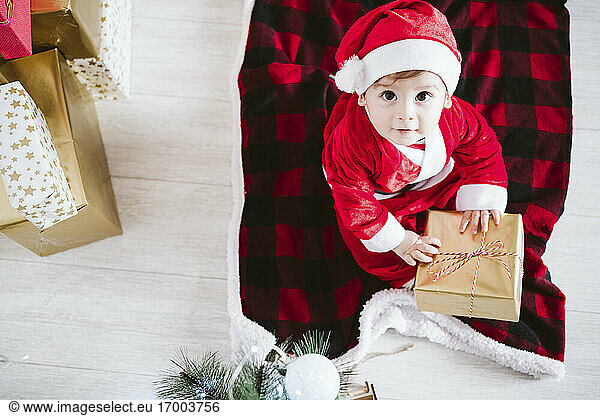 Cute baby boy in Santa Claus costume playing with gift box while sitting at home during Christmas