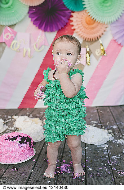 Cute baby girl eating birthday cake while standing on floorboard