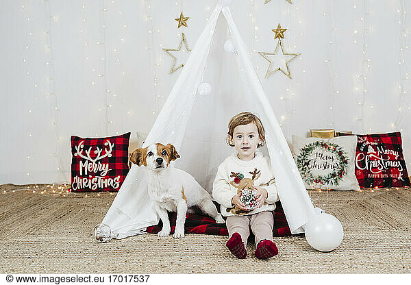 Cute baby girl with puppy sitting in tent against Christmas decorations at home