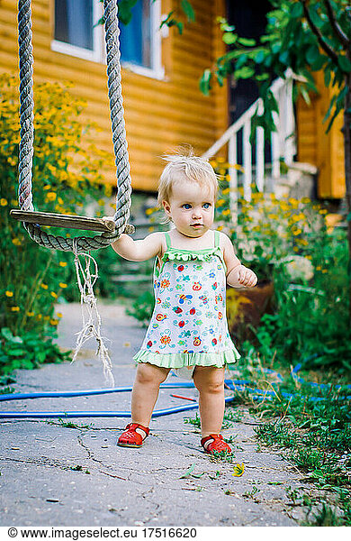 Cute baby girl 3-4 year old in the garden plays a rustic swing
