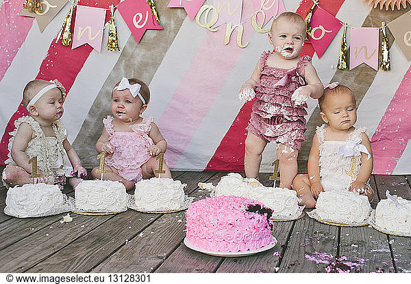Cute baby girls with birthday cakes on floorboard against decoration at party