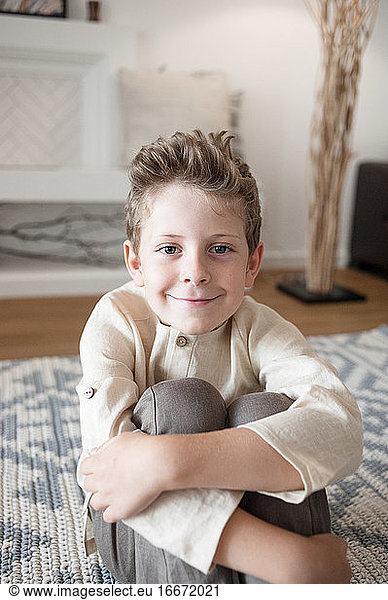 Cute boy in linen home clothes looks at a camera sitting on floor.