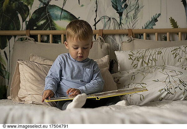 Cute boy reading book sitting on bed at home