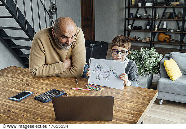 Cute boy showing drawing through laptop sitting by grandfather at home