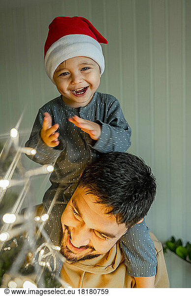 Cute boy sitting on father's shoulder decorating Christmas tree at home