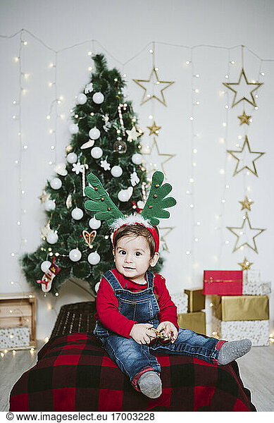 Cute boy wearing horned headband sitting against Christmas tree at home during Christmas