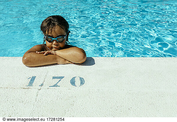 Cute boy with arms crossed leaning at poolside on sunny day