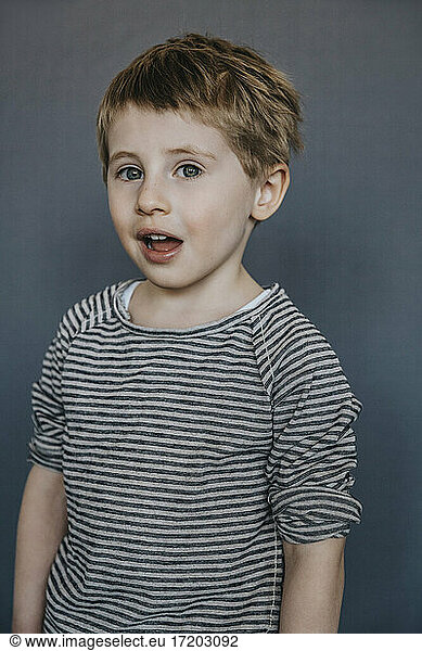 Cute boy with mouth open standing against gray background